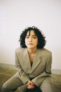 Photo of Duygu Agal taken from a slightly elevated position: She*He has shoulder-length, curly dark hair, wears a beige trouser suit and several thin silver necklaces. She*He is casually seated, leaning slightly forward.