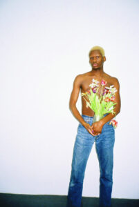 Ein junger Schwarzer Mann mit kurzen blondierten Haaren steht oberkörperfrei in Jeans vor einer weißen Wand. Er hält eine Vase mit einem Strauß Tulpen vor sich und schaut in die Kamera.