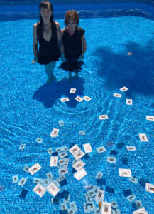 Little Annie and Evilyn Frantic stand drenched in black dresses close together in a turquoise pool, a multitude of playing cards floating in the water in front of them.