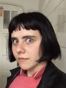 A young white woman with a black bob haircut looks sideways into the camera with wide eyes. She is wearing a pink wool sweater and a black jacket. Behind her is a white tiled wall and a white wooden door.