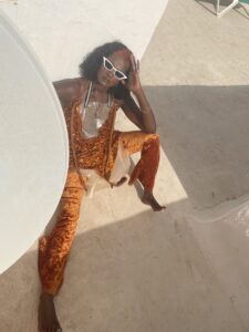 Ein Foto von schräg oben von einer Schwarzen Frau mit dunklen, offenen Locken auf Schulterlänge sitzt barfuß auf einem sonnenbestrahlten Betonboden. Sie trägt kunstvolle Kleidung aus orangenem Samt und mattem Plastik sowie eine weiße Sonnenbrille.