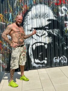 Eine muskulöse Person mit vielen Tattoos steht oberkörperfrei vor einer Grafitti-Wand. Sie trägt eine knielange Hose und hellgrüne Sportschuhe. Sie hat einen grauen Bart und trägft eine Glatze