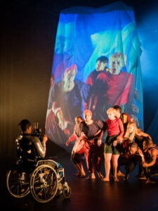 Eine Person im Rollstuhl vorn auf der Bühne und daneben eine Gruppe eng beieinander stehender Tänzer*innen vor einer Leinwand.