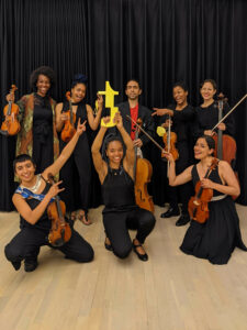 The eight musicians of String Archestra, seven women and one man, all wear black clothes, smile joyfully into the camera, their string instruments in their hands. A female musician in the middle holds up a prize or trophy.