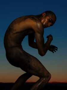 An artistic photographic work of a naked, black tattooed man. He kneels slightly, his upper body bent rigidly into a hollow circle, his arms loosely folded and looking sideways at the camera. In the background a blue-pink evening sky.
