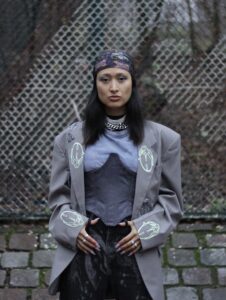 Maque Tumai wears a stylish combination of shirt, satin corsage and wide jacket, in addition a floral bandana, her hands casually placed on her belt.