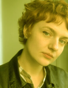 Greenish cast photo of a young white woman with short wavy hair. She tilts her head and looks slightly sideways at the camera.