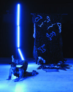 On a blue-lit stage, a man with a moustache, durag and patterned trousers lies on his stomach, his chest lifted, one leg twisted back and up. Behind him are two objects made of neon tubes, braided fabric and fur.