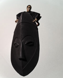 Vor einem weißen Hintergrund schwebt eine große Schwarze Skulptur, welche wie eine Maske oder ein Gesicht aussieht. Oben, hinter der Skulptur sieht man die obere Körperhälfte einer Frau in schwarzer Kleidung. Sie hat hellblaue Farbe unter den Augen.