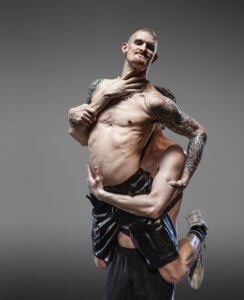 A white, thin, heavily tattooed man with very short hair is being lifted up by another man. Both are topless and wearing denim shorts and trainers. The man being lifted holds on to the other's arms and grins.