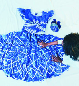 Auf einem weißen Boden liegt ein strahlend blaues Kleid, kunstvoll bestickt mit weißen Streifen im Zickzackmuster. Eine Schwarze Frau streicht mit ihren Händen über das Kleid.