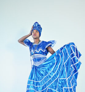 Eine schlanke Schwarze Frau trägt ein strahlend blaues, kunstvolles Kleid mit weißem Muster und einen Turban im passenden Stoff. Sie hält den weiten Rock mit einer Hand seitlich hoch. Die andere Hand hält sie an ihren Kopf und blickt nach oben.