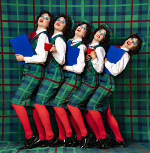 Fünf Frauen in grünen schottischen Tartan-Kleidern, weißen Blusen, roten Strumpfhosen und knallblauem Lidschatten stehen eng in einer Reihe vor einem Hintergrund im Muster wie ihre Kleider. Sie halten blaue Klemmbretter und rote Tassen in den Händen.