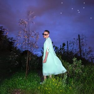 Michael Brailey has short, light brown hair, wears a loose, light blue dress, black sunglasses and looks quirkily over the shoulder. In the background, an artificial-looking meadow and trees, purple-blue night sky.