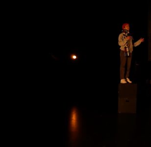 A white person stands on a black pedestal on a dark stage. With a mobile phone in her right hand and her mouth open. She is wearing grey trousers and a jumper, a red cap, a colourful scarf and white trainers. Her left arm is raised