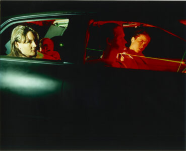 Lateral view of the four occupants of a car, whose faces are illuminated in the interior in green at the back and red at the front. Two young faces are clearly visible. An older woman sits on the back seat in the dark with her mouth agape.