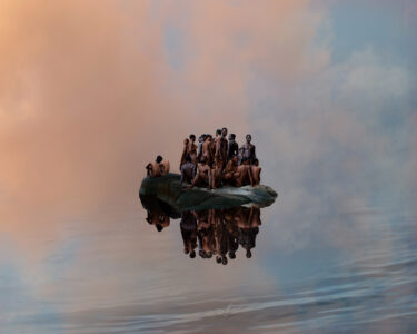 An artistic photographic work showing a group of black naked people standing and squatting on a large rock. The rock floats in front of a pink cloud facade, waves are indicated on the lower right. The people are reflected below the rock.