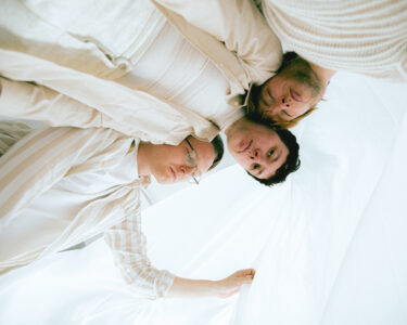A photo of three young white people with short hair and creamy white clothes photographed from below. They hold a white cloth over their heads and lean tenderly against each other.