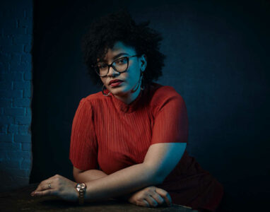 A Black woman with a natural Afro, glasses, red lipstick and a red T-shirt sits with her arms crossed in front of a dark background. Her hands rest on her knees, she looks calmly and seriously into the camera with a slightly tilted head.