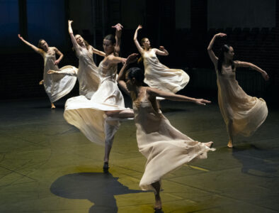 Six dancers in white dresses move swingingly on a greenish-lit stage floor. They swing their arms and legs up and to the side. Their dresses are made of delicate fabric, their skirts swing around airily.