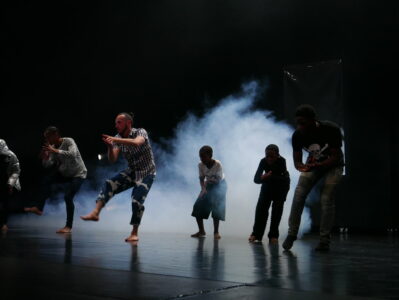 Fünf Tänzer*innen, davon zwei Kinder, stehen auf jeweils einem Bein in einem dunklen Raum, der von hinten mit weißem Nebel erfüllt ist. Sie klatschen in die Hände.