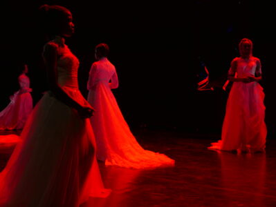 In a dark, red-lit room, four women in wedding dresses walk around sublimely. The light makes the dresses look blood-red.