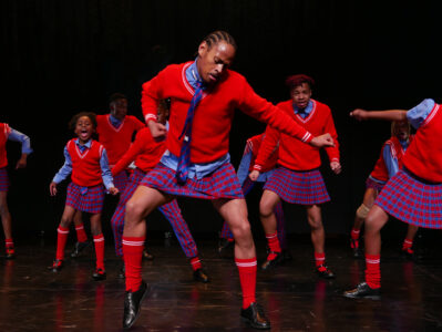 A group of Black dancers consisting of adults and children are spread out in a dark stage space. They wear red jumpers, red and blue checked skirts and trousers and red stockings and move tensely.