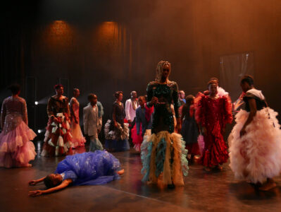 Fourteen black adults and children in pompous costumes with tulle skirts, frills and glitter stand with their backs to the camera in a dark stage space. A child in blue lies backwards on the floor, hands outstretched above her head.