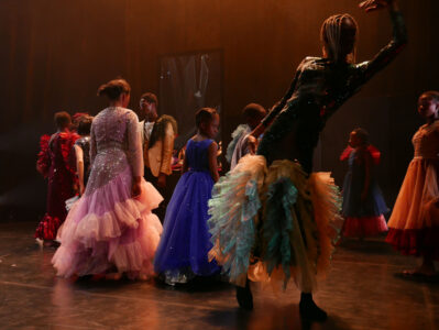 Ten Black adults and children in pompous costumes with tulle skirts, frills and glitter stand with their backs to the camera in a dark stage space. One person in front moves elegantly in a dance pose.