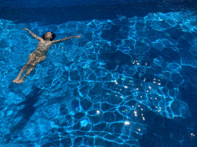 Evilyn Frantic floats naked in a turquoise pool on her back, her arms stretched out to her sides.