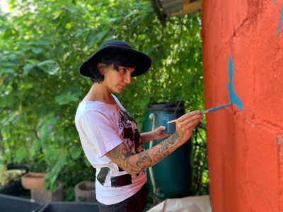 Eine Frau mit weißem T-Shirt und blauem Hut malt mit einem Pinsel einen blauen Vogel auf eine orangene Wand und schaut konzentriert auf ihr Werk. Hinter ihr wächst dichtes grünes Gebüsch.
