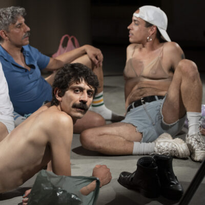 Four people on a stage floor. Two of them in the background are wearing shorts, one a shirt, the other a bra and a cap. They are talking. The person in front is naked and turns to face the camera lying down. Another one is half out of the picture.