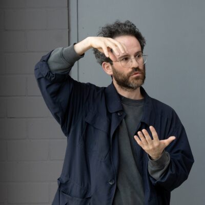 Eine männlich gelesene Person in einem grauen Pullover und schwarzen Hemd hält beide Hände in einer Haltung, als würden sie eine Kugel halten.