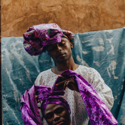 Zwei Schwarze Personen in leichter, lockerer Kleidung mit Mustern und lila Tüchern um den Kopf stehen hintereinander. Die Hintere Person bindet liebevoll das Tuch der vorderen um deren Kopf. Sie haben ihre Augen sanft geschlossen.