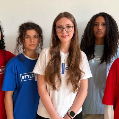 Five people with different hair lengths and textures are wearing colorful T-shirts with the words “STYLE CREATE” printed on them and are standing in front of a light-colored wall.
