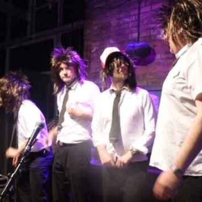 Fünf Erwachsene mit weißen Hemden und Krawatten stehen in einem Raum mit Backsteinwänden und bereiten sich auf eine musikalische Darbietung vor.