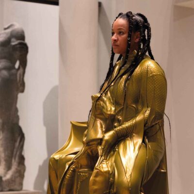 Eine Person in einen glänzenden goldfarbenen Anzug sitzt als Ausstellungsstück im Museum.