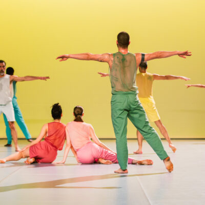 5 Tänzer*innen in bunten Kostümen stehen mit ausgestreckten Armen vor einer gelben Wand. Zwei Tänzer*innen in rosa und roten Kostümen sitzen in der Mitte des Raumes aneinander gelehnt auf dem weißen Boden.