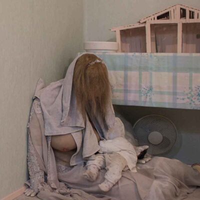 Eine Person mit langen Haaren sitzt mit einem Baby auf dem Boden neben einem Tisch mit einem Modellhaus.
