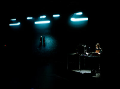 In einem dunklen Bühnenraums steht eine Person vor einem Tisch mit vielen elektronischen Musikgeräten und Computern. Daneben wird ins Dunkle ein Bild von zwei wandelnden Figuren projiziert.