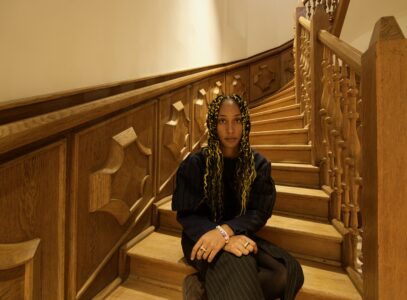 Isocialbutterflyy wears rasta braids with yellow strands woven into them, a black jumper and a black pinstripe skirt. She sits casually on a stiff of a wood-panelled stairwell.