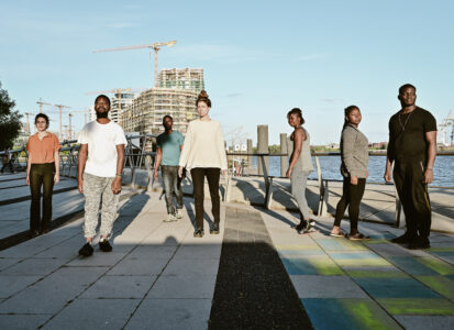 Seven people with different skin colours stand on a square at Hamburg harbour and are illuminated by the sun.