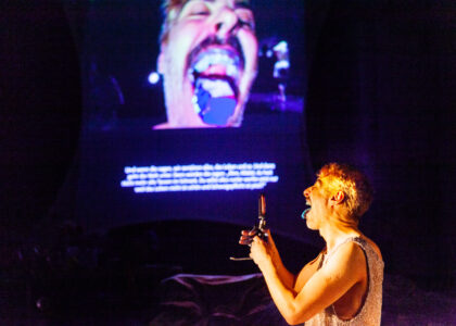 A performer steckt seine Zunge raus in eine Handy Kamera. Sein Gesicht mit aufgerissenem Mund und rausgestreckter blauer Zunge ist groß auf einer Leinwand projiziert.