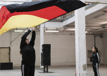 Eine blonde Frau im schwarzen Addidas-Trainingsanzug und weißen Handschuhen schwenkt eine riesige Deutschlandfahne über sich. Eine zweite Frau im gleichen Outfit steht an einer Säule im rechten Bildrand. Im Hintergrund setehen Musikboxen