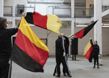 Four people in black Addidas tracksuits and white gloves wave huge German flags around them. A man and a woman stand close to each other in the middle. Another man and a woman stand further away and look at them