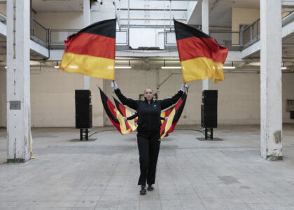 In a bright hall with white bars, people in black tracksuits wave German flags.