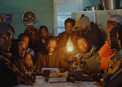 Some people of the village community from young to old are sitting around a table, looking tense and concentrated. The room is lit by an oil lamp on the table, next to it a plate with bread.