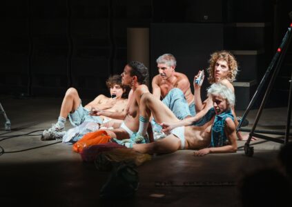 5 people sit and lie on the floor in a relaxed posture. Some support themselves on the floor. They are lightly clothed. One person holds a small bottle in his hand and looks into the camera.