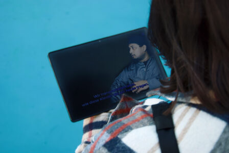 Eine Person vor einem blauen Hintergrund schaut auf ein Tablet, welches sie in den Händen hält. Auf dem Tablet läuft ein Video, welches einen sprechenden Mann in blauem Overall und einer blauen Kappe zeigt.