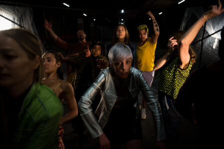 Eight diverse performers in colorful costumes jump towards the camera in a dark room. They throw their arms forward, up and down, creating dynamics in the image. The face of the person in front is cut off from the left edge of the image.